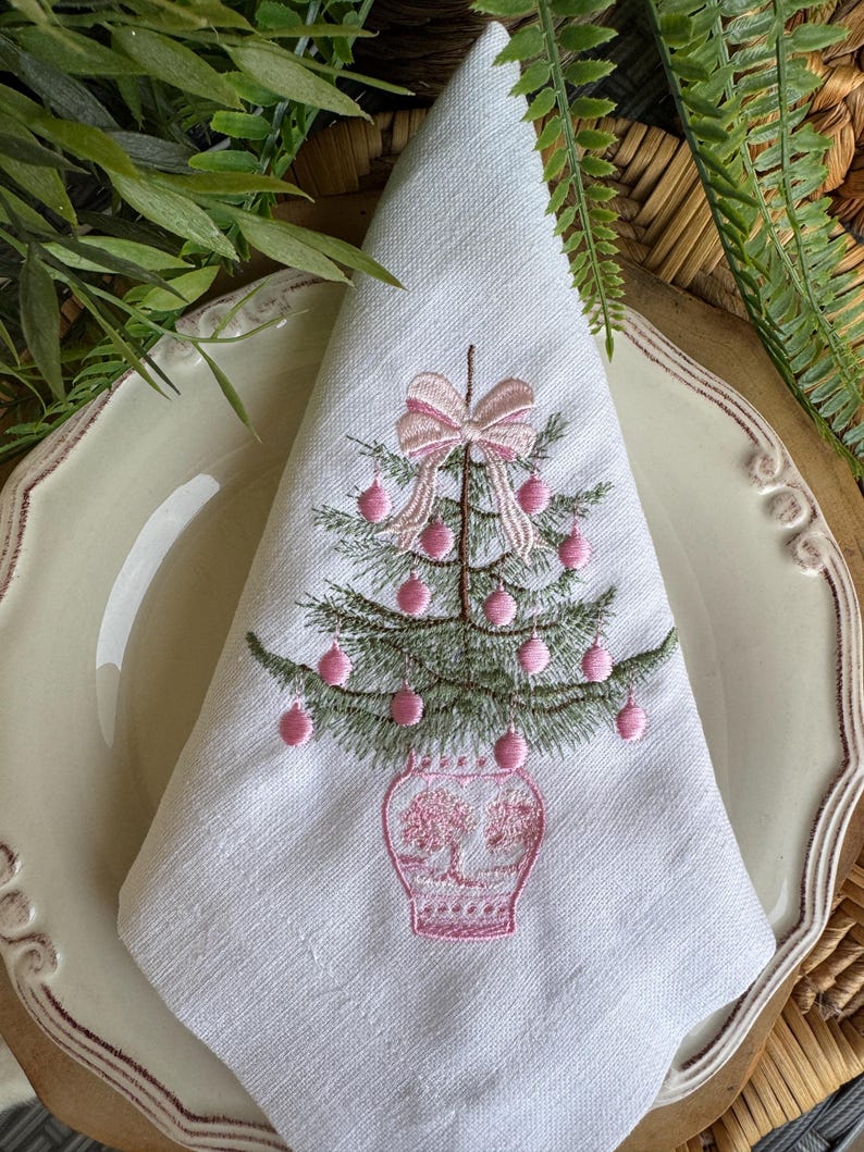 May include: White linen napkin featuring an embroidered Christmas tree design. The tree is green with pink baubles and a pink bow. The tree sits in a pink vase. The napkin is folded and placed on a white plate with a decorative edge.
