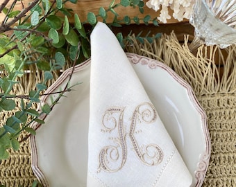 May include: A white linen napkin with a delicate embroidered letter "K" in a beige color. The napkin is folded and placed on a white plate with a pink rim. The plate is on a woven placemat.
