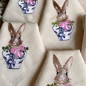 May include: A set of cream-colored cloth napkins with embroidered bunny designs. Each napkin features a brown bunny in a blue and white vase with a pink bow. The napkins are folded and arranged for display.