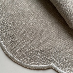 Puede incluir: Un mantel individual redondo de lino beige con un borde festoneado y costuras blancas. El mantel individual est&aacute; doblado por la mitad, mostrando la textura de la tela.