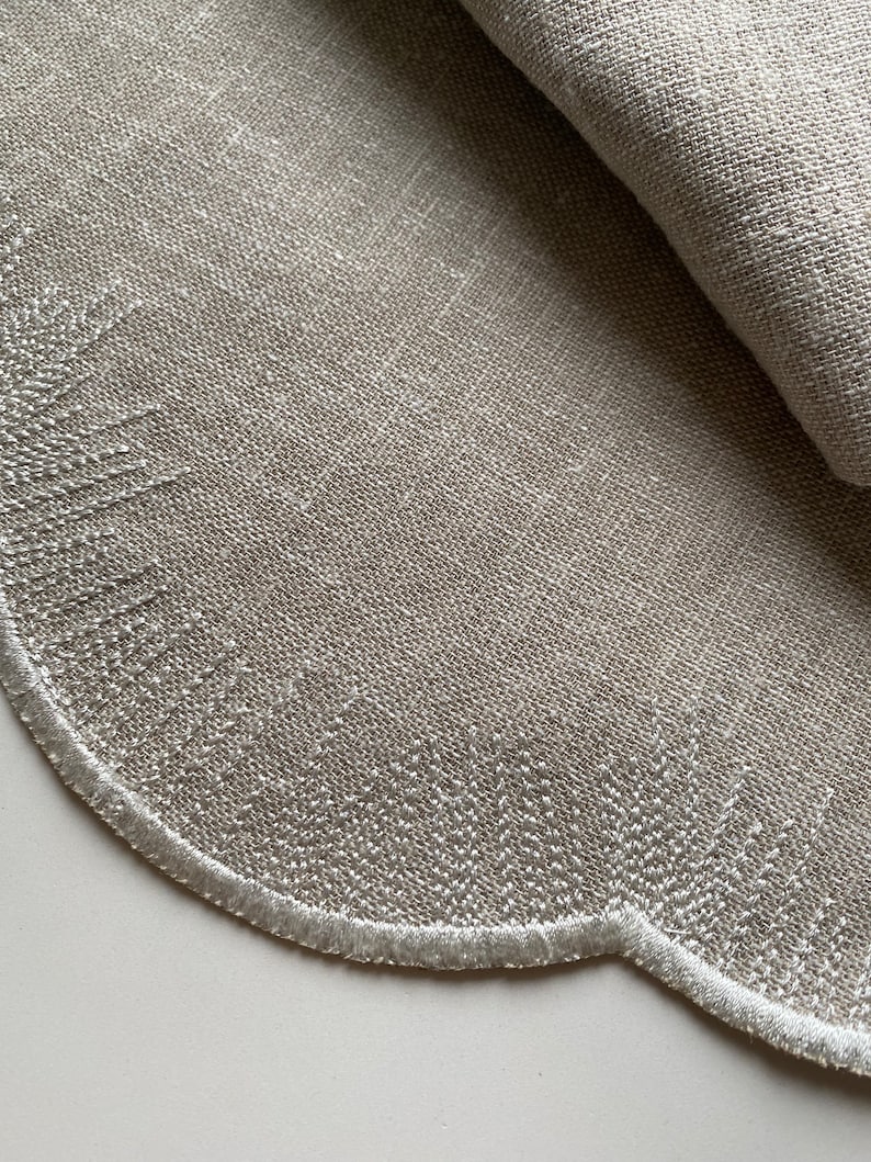 Puede incluir: Un mantel individual redondo de lino beige con un borde festoneado y costuras blancas. El mantel individual est&aacute; doblado por la mitad, mostrando la textura de la tela.