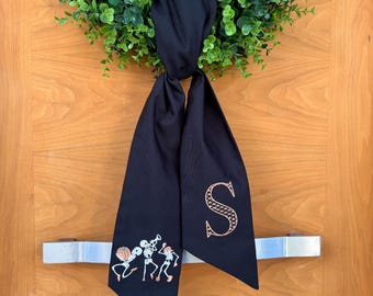 May include: A black fabric door wreath with a gold embroidered letter "S" and a design of three skeletons playing musical instruments.
