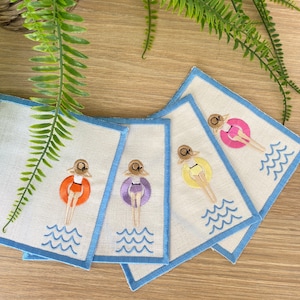 Peut inclure: Cinq dessous-de-verre en lin brodés avec une bordure bleue. Chaque dessous-de-verre présente un maillot de bain et une bouée de couleur différente avec une femme à l'intérieur. Les dessous-de-verre sont disposés en éventail sur une surface en bois.