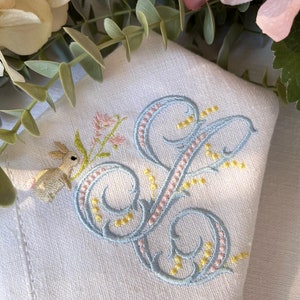 May include: A white linen napkin with a blue and pink embroidered design. The design features a stylised letter "S" with a small bunny and flowers.