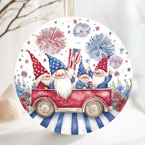 May include: Round, white, wooden ornament with a red, white, and blue illustration of four gnomes riding in a red car with an American flag. The gnomes are wearing red, white, and blue hats. The background is white with red, white, and blue fireworks.