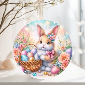 Puede incluir: Adorno de Pascua redondo con una ilustración en acuarela de un conejito con una corona floral, sosteniendo una cesta de huevos decorados. El adorno está adornado con un lazo rosa y rodeado de flores en colores pastel. Se adjunta una cuerda para colgar.