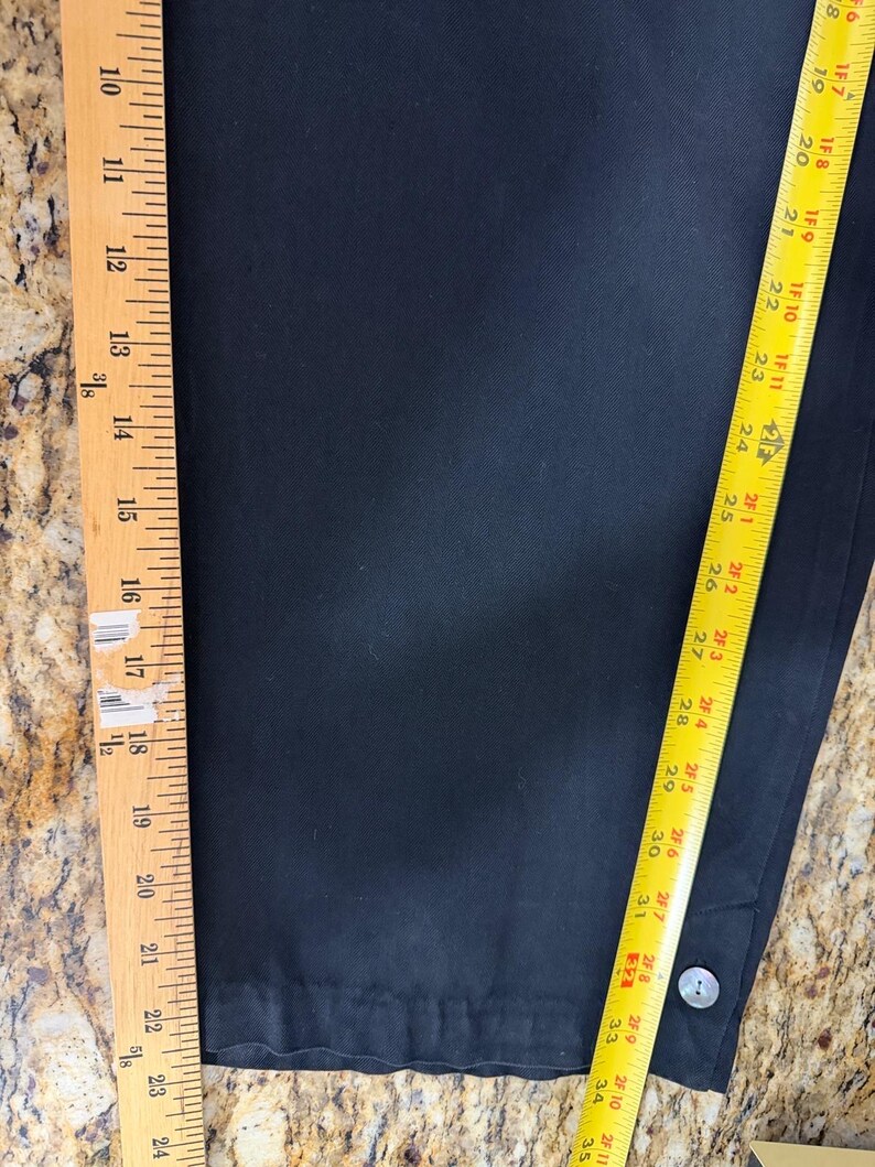 K&ouml;nnte beinhalten: Schwarze Hose mit Knopfdetail. Das Kleidungsst&uuml;ck wird mit einem Lineal gemessen und zeigt eine L&auml;nge von etwa 86 cm. Der Stoff scheint eine einfarbige, dunkle Farbe zu haben.