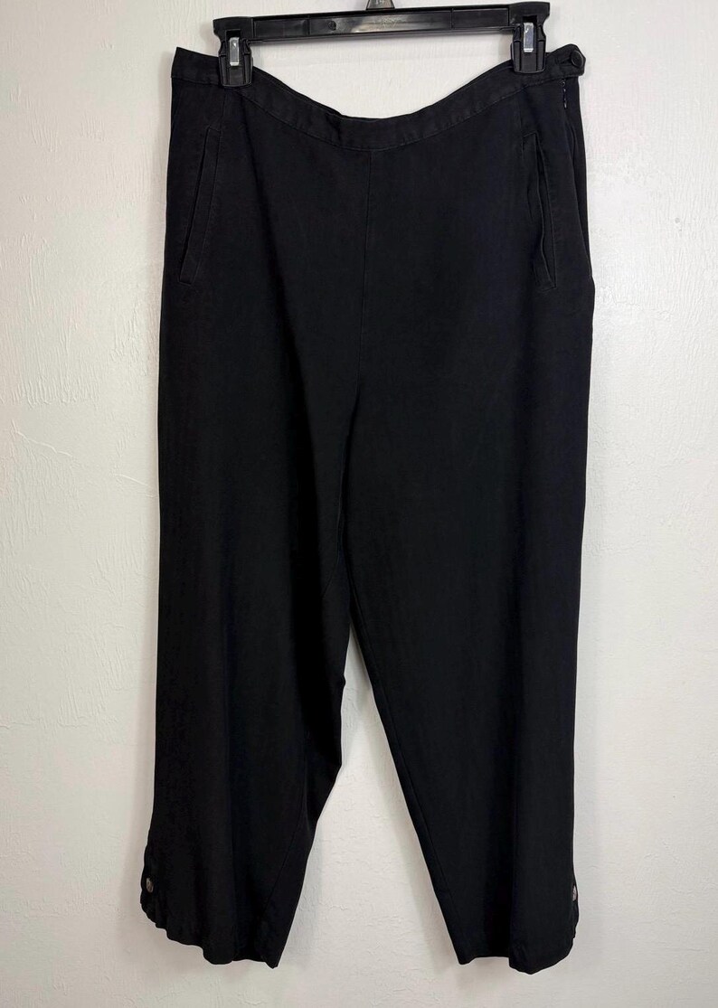 K&ouml;nnte beinhalten: Schwarze, knielange Hose mit hoher Taille und Seitentaschen. Die Hose hat ein Knopfdetail am Saum und h&auml;ngt an einem schwarzen Kleiderb&uuml;gel. Der Stoff scheint ein leichtes Material zu sein.