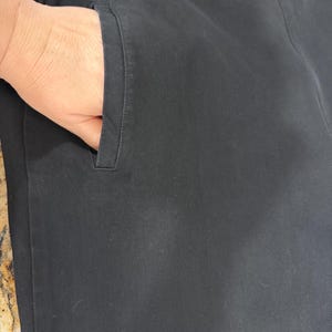 K&ouml;nnte beinhalten: Nahaufnahme einer schwarzen Hose mit einer Hand in der Tasche. Die Hose ist aus einem einfarbigen, dunklen Stoff gefertigt. Die Tasche ist schlicht und zweckm&auml;&szlig;ig gestaltet.
