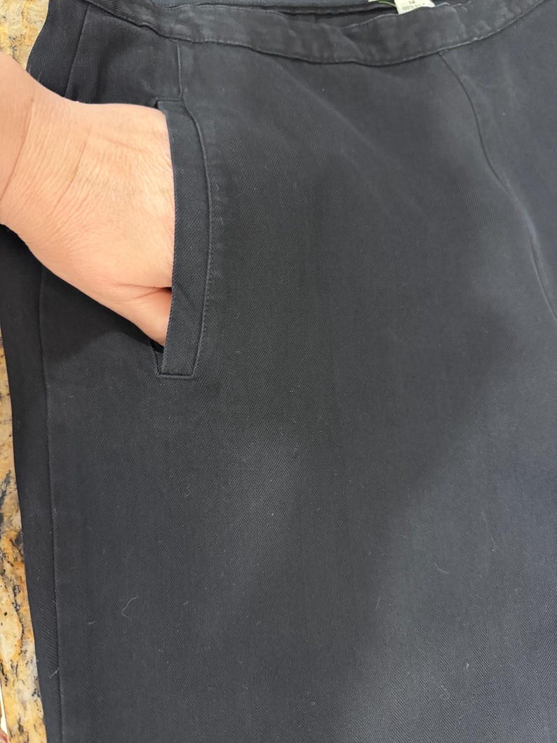 K&ouml;nnte beinhalten: Nahaufnahme einer schwarzen Hose mit einer Hand in der Tasche. Die Hose ist aus einem einfarbigen, dunklen Stoff gefertigt. Die Tasche ist schlicht und zweckm&auml;&szlig;ig gestaltet.