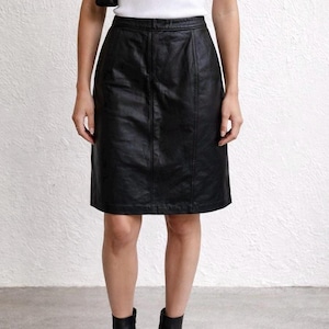 Puede incluir: Falda de cuero negra con dobladillo a la altura de la rodilla. La falda tiene un diseño clásico y entallado con costuras verticales y cintura ajustada. La modelo lleva botines negros.