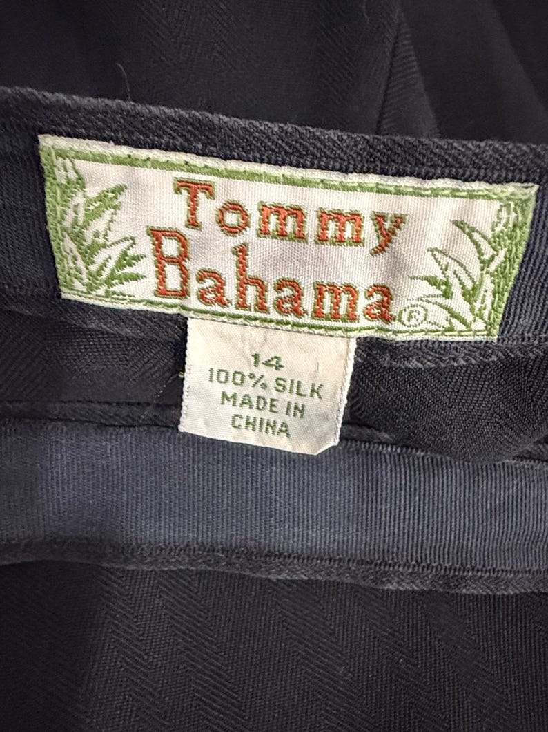 K&ouml;nnte beinhalten: Nahaufnahme eines marineblauen Kleidungsst&uuml;cketiketts mit dem Markennamen "Tommy Bahama" in orangefarbenen Buchstaben. Das Etikett gibt au&szlig;erdem an, dass das Kleidungsst&uuml;ck Gr&ouml;&szlig;e 14 hat und aus 100 % Seide besteht, hergestellt in China.