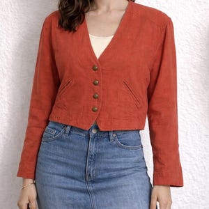 May include: A burnt orange blazer with long sleeves and a V-neckline. The blazer has four gold-colored buttons and two small pockets. The model is wearing a white tank top and a denim skirt.