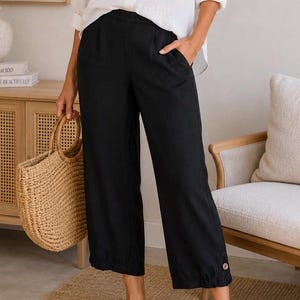 K&ouml;nnte beinhalten: Schwarze, cropped Hose mit Knopfdetail am Saum. Die Hose wird mit einem wei&szlig;en Hemd und schwarzen Sandalen kombiniert. Eine gewebte Tragetasche wird gehalten. Das Outfit ist l&auml;ssig und f&uuml;r warmes Wetter geeignet.
