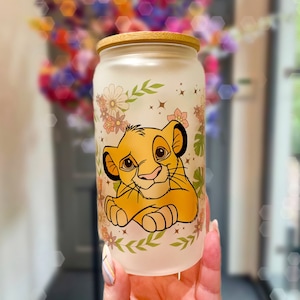 Könnte beinhalten: Ein gefrosteter Glasbecher in Dosenform mit einem lächelnden Simba aus Der König der Löwen. Der Becher ist mit floralen und Blattakzenten verziert und hat einen Bambusdeckel. Das Design ist in Gelb-, Orange- und Grüntönen gehalten.