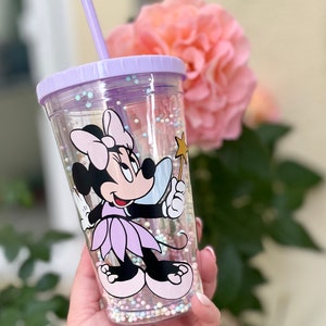 Könnte beinhalten: Ein durchsichtiger Kunststoffbecher mit lila Deckel und Strohhalm, mit einem Minnie Mouse-Design. Der Becher ist mit kleinen, bunten Perlen gefüllt und hat ein Cartoon-Bild von Minnie Mouse, die einen Sternenstab hält. Im Hintergrund eine verschwommene rosa Blume.