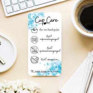 Könnte beinhalten: Eine weiße Karte mit dem Text "Cup Care" und Pflegehinweisen auf Deutsch. Die Karte zeigt blaue Aquarellakzente und Symbole, die nur Handwäsche, nicht mikrowellengeeignet, nicht spülmaschinengeeignet und nicht einweichen anzeigen.