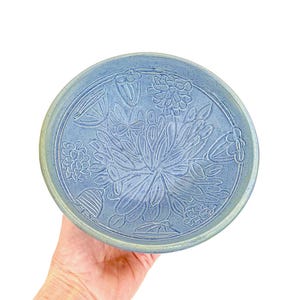 May include: A round, light blue ceramic plate with a floral design. The plate features an embossed pattern of flowers and leaves. The rim has a light green hue. The plate is held by a hand against a white background.