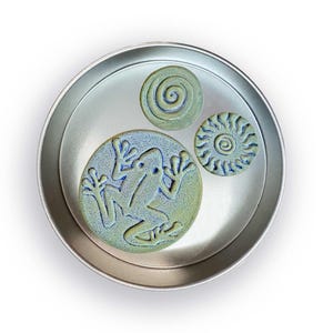 May include: A circular, silver metal tray contains three ceramic pieces. The largest piece features a frog design in blue and green. Two smaller pieces have spiral and sunburst patterns. The ceramic pieces have a textured appearance.