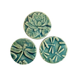 May include: Three teal ceramic tiles with nature-themed designs. One tile depicts a lotus flower, another a dragonfly, and the third features dragonflies. The tiles have a glossy finish and are set against a white background.