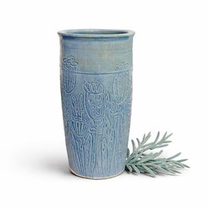 May include: A tall, light blue ceramic vase with embossed floral and plant designs. The vase has a cylindrical shape and a slightly textured surface. A sprig of gray-green foliage rests beside the vase.