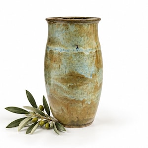 May include: A cylindrical ceramic vase with a flared rim, featuring a mottled glaze in shades of earthy brown, tan, and muted blue. A small sprig of green olive leaves with several small green olives is positioned at the base of the vase.