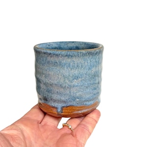 May include: A handmade ceramic tumbler with a textured, blue and brown glaze. The cup has a cylindrical shape and is held in a hand. The blue glaze covers the majority of the cup, with a brown band at the base.