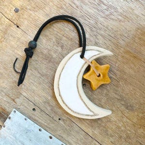 May include: A white ceramic crescent moon with a yellow ceramic star hanging from a black cord. The moon and star are hanging on a wooden surface.