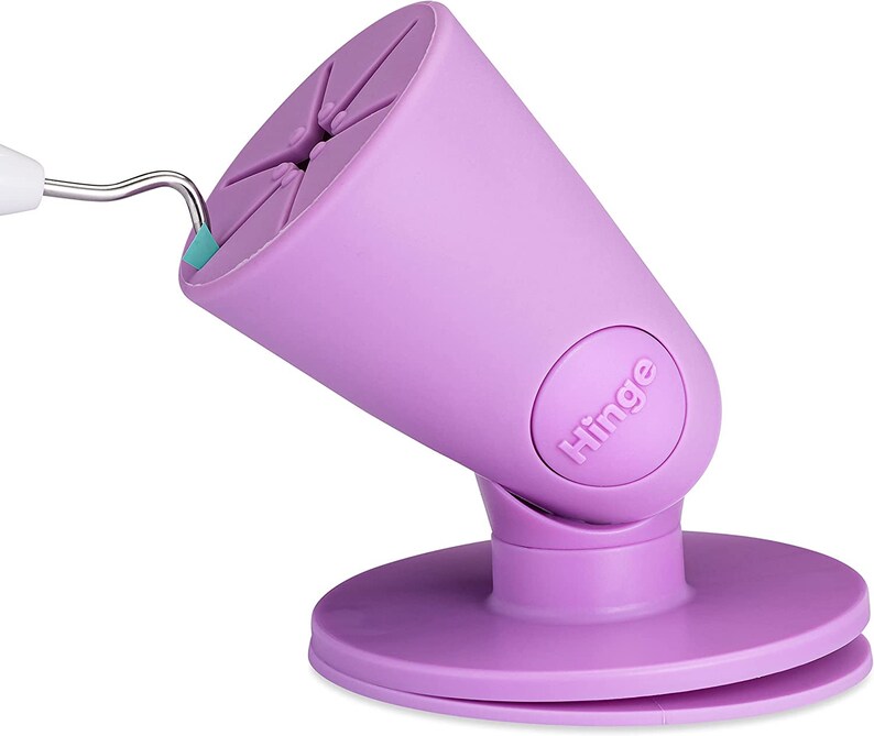 Puede incluir: Soporte para tel&eacute;fono morado con base de ventosa y bisagra. El soporte est&aacute; dise&ntilde;ado para sostener un tel&eacute;fono en posici&oacute;n vertical. El soporte tiene la palabra "Hinge" impresa en &eacute;l.