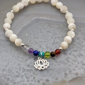 Könnte beinhalten: Armband aus weißen Edelsteinen mit silbernen Perlen und einem silbernen Lotus-Anhänger. Das Armband ist mit sieben kleineren Edelsteinen in Regenbogenfarben verziert.
