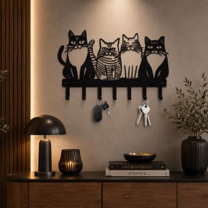 Può includere: Un portachiavi da parete in metallo nero con il design a silhouette di quattro gatti seduti in fila. La barra orizzontale include sei ganci rivolti verso il basso, con due mazzi di chiavi in metallo appesi ai ganci su una parete neutra.