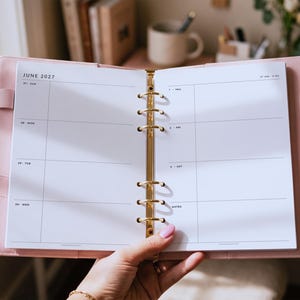 May include: A light pink six-ring binder planner open to a weekly spread for June 2027. The white pages feature black text with days of the week and a notes section. A gold-toned metal ring mechanism secures the pages in the center of the cover.