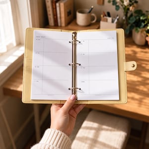 May include: A light yellow three-ring binder planner open to a weekly calendar page for July 2027. The white paper features black text with days of the week from Monday to Sunday and a notes section. The binder has a metal ring mechanism.