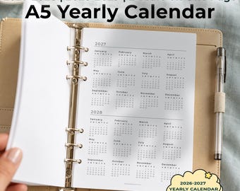 Op de afbeelding: Een A5-jaarkalender voor een ringband, met 2027 en 2028 op één pagina. Het witte papier bevat zwarte tekst en maandoverzichten. Een doorzichtige pen zit in een beige penlus naast de open pagina's van de ringband.