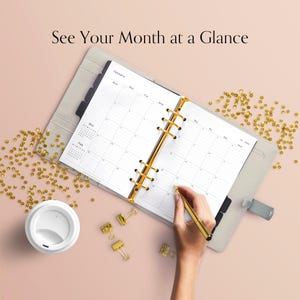 May include: A light grey planner open to a January calendar page, with a gold pen in hand, and the text "See Your Month at a Glance". The planner has gold accents. A white coffee cup and gold paper clips are also visible.