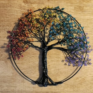 May include: A handcrafted wire tree sculpture with a circular frame. The tree features a black twisted trunk and branches, with colorful beaded leaves in shades of red, orange, yellow, green, blue, and purple. The artwork is displayed on a wooden surface.