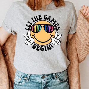 May include: A gray t-shirt with a yellow smiley face wearing sunglasses that say "Field Day". The text "Let the Games Begin!" is written around the smiley face.