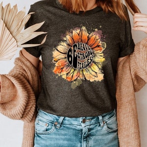 May include: A dark gray t-shirt with a sunflower graphic and the text "Brave Strong Fearless" inside the sunflower. The sunflower is painted in shades of orange and yellow.