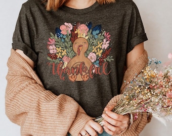 May include: A dark gray t-shirt with a colorful illustration of a turkey surrounded by flowers and the text "Thankful". The model is wearing the shirt with a brown cardigan and blue jeans.