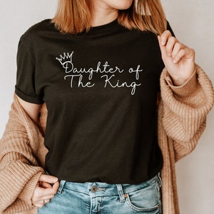 Puede incluir: Una camiseta negra con una corona blanca y el texto "Daughter of The King" impreso en la parte delantera.