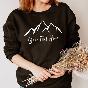 以下が含まれることがあります： 白い山と「Your Text Here」の文字がプリントされた黒いスウェットシャツ