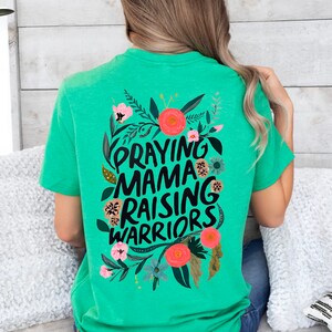 Puede incluir: Camiseta verde con un dise&ntilde;o floral y el texto "Praying Mama Raising Warriors" en negro.