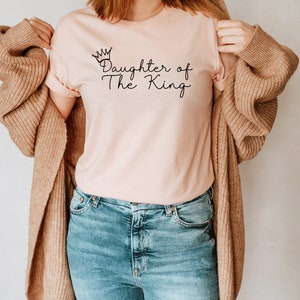 Puede incluir: Una camiseta rosa con el texto "Daughter of The King" y un gr&aacute;fico de corona.