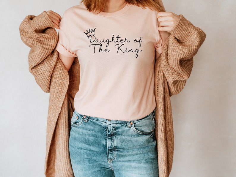 Puede incluir: Una camiseta rosa con el texto "Daughter of The King" y un gr&aacute;fico de corona.