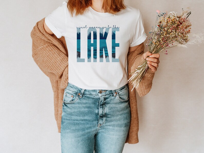 K&ouml;nnte beinhalten: Ein wei&szlig;es T-Shirt mit einer blauen und wei&szlig;en Grafik, auf der "I just wanna go to the LAKE" steht. Die Grafik zeigt eine Seenszene mit B&auml;umen und Wasser.