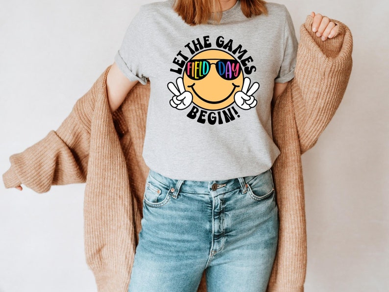 May include: A gray t-shirt with a yellow smiley face wearing sunglasses that say "Field Day". The text "Let the Games Begin!" is written around the smiley face.