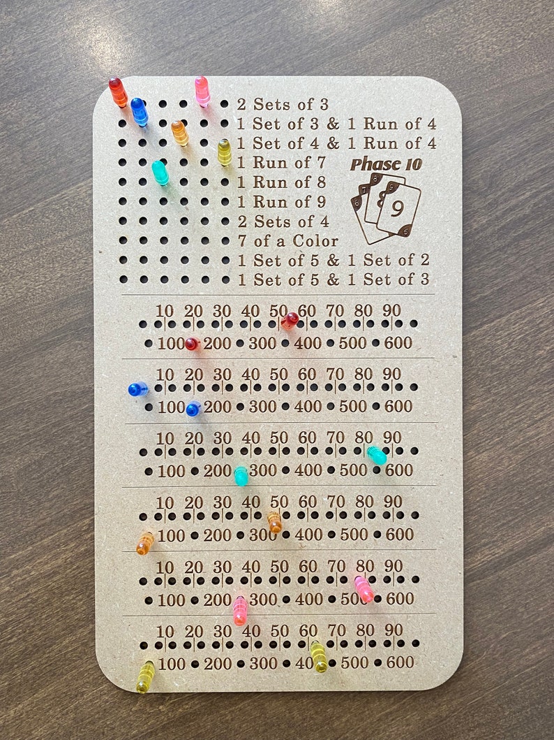 May include: A wooden game board with a grid of holes and pegs. The board has a list of scoring rules for the game Phase 10, including sets of 3, runs of 4, and sets of 5. The board also has a number scale from 10 to 600.