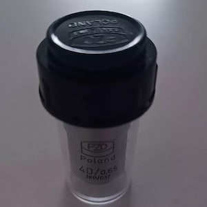 May include: A clear glass vial with a black cap. The vial has the text "PZO Poland" and measurements of 40/0.65 and 160/0.17 printed on it. The vial is cylindrical and appears to be empty.