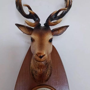 May include: A wooden wall-mounted barometer with a deer head sculpture. The barometer has a white face with black markings and the text "VARIABLE LYMAN-DALLILA BAROMETRE PARIS" and "TRES SEC".