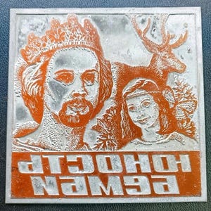 Puede incluir: Impresión en relieve metálico cuadrado con un diseño naranja y plateado. La imagen presenta un hombre coronado, una mujer y un ciervo. El texto en la parte inferior dice "EWEN HOCLIP".